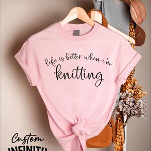 Pu&ograve; includere: T-shirt rosa chiaro con la scritta "life is better when I'm knitting" in corsivo nero. La maglietta &egrave; annodata in basso e appesa a una gruccia di legno. Sono visibili anche le parole "Custom Infinity Gifts".