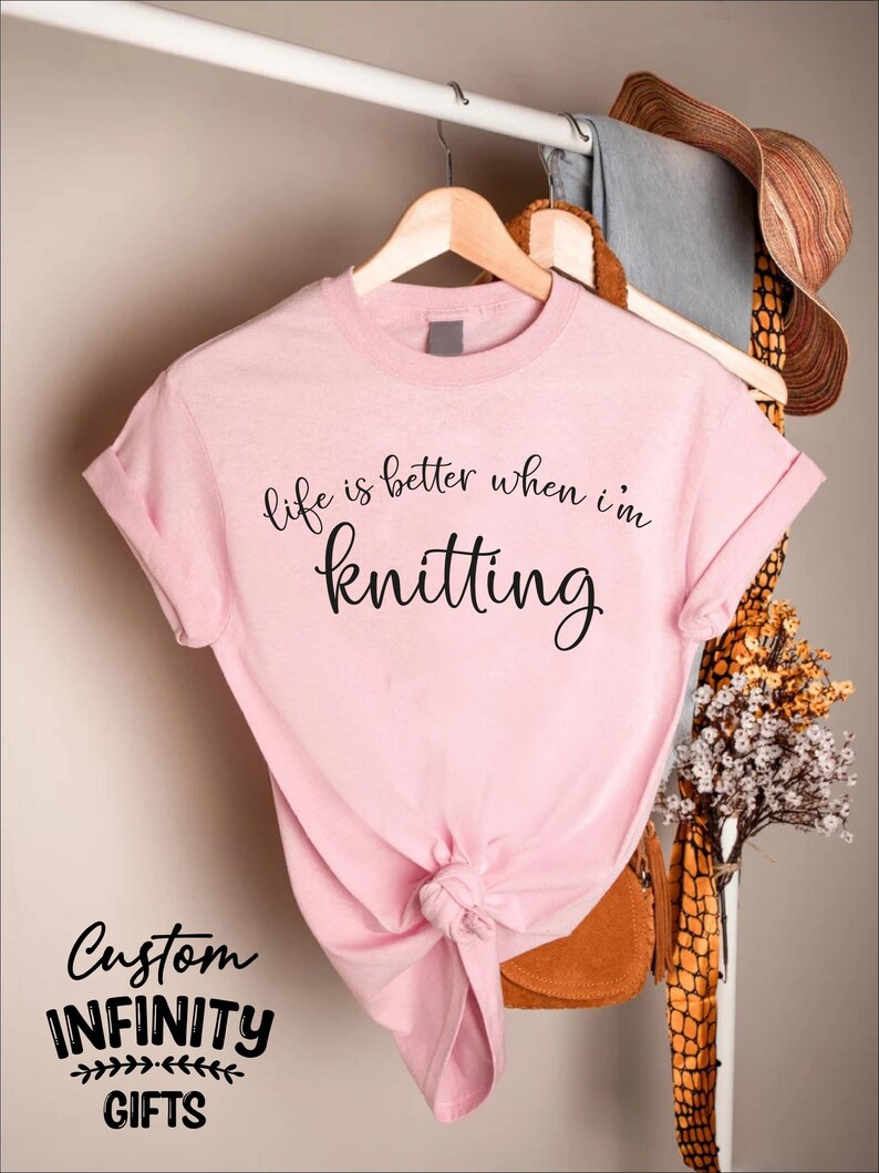 Pu&ograve; includere: T-shirt rosa chiaro con la scritta "life is better when I'm knitting" in corsivo nero. La maglietta &egrave; annodata in basso e appesa a una gruccia di legno. Sono visibili anche le parole "Custom Infinity Gifts".