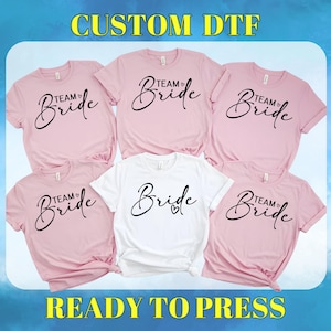 May include: Five iron-on transfer designs for t-shirts. The designs are pink and white and feature the text "Team Bride" in a cursive font.