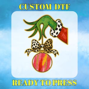May include: A green hand with a red sleeve and a black and white polka dot cuff holds a leopard print bow and a red and gold ornament. The text "CUSTOM DTF" and "READY TO PRESS" are in yellow.