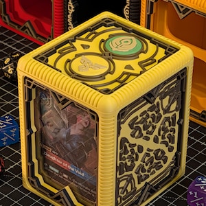 May include: A yellow cube-shaped card holder with black decorative accents and a card visible through the front. The card features the text "Daughter of the Void". Other similar boxes are visible in the background.