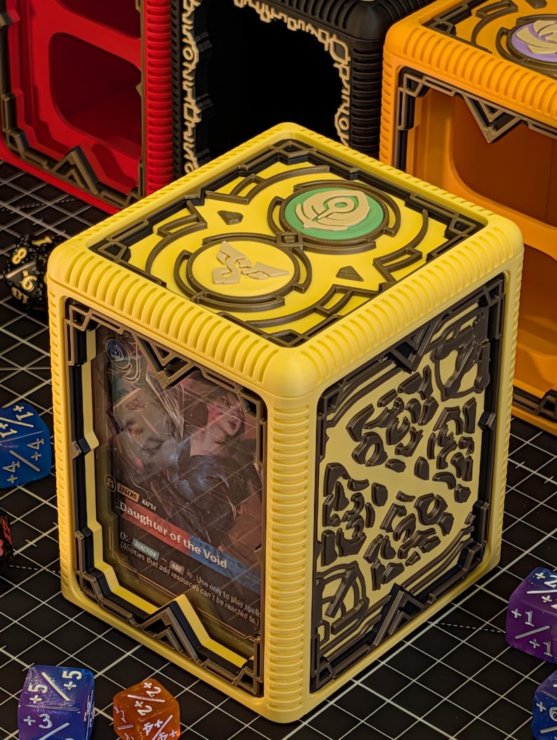 May include: A yellow cube-shaped card holder with black decorative accents and a card visible through the front. The card features the text "Daughter of the Void". Other similar boxes are visible in the background.