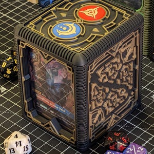 May include: Black and gold card storage box with intricate geometric designs. The top features two circular emblems in red and blue. A card with the text "Daughter of the Void" is visible inside. Dice of various colors surround the box.