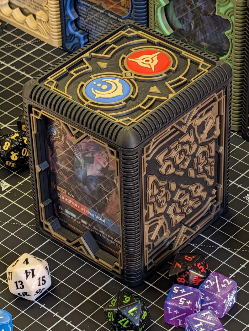 May include: Black and gold card storage box with intricate geometric designs. The top features two circular emblems in red and blue. A card with the text "Daughter of the Void" is visible inside. Dice of various colors surround the box.