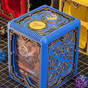 May include: A blue and gold card storage box with intricate geometric designs. The box features a card with the text "Daughter of the Void." Other boxes in red, black, and yellow are visible in the background. Dice are scattered around the box.