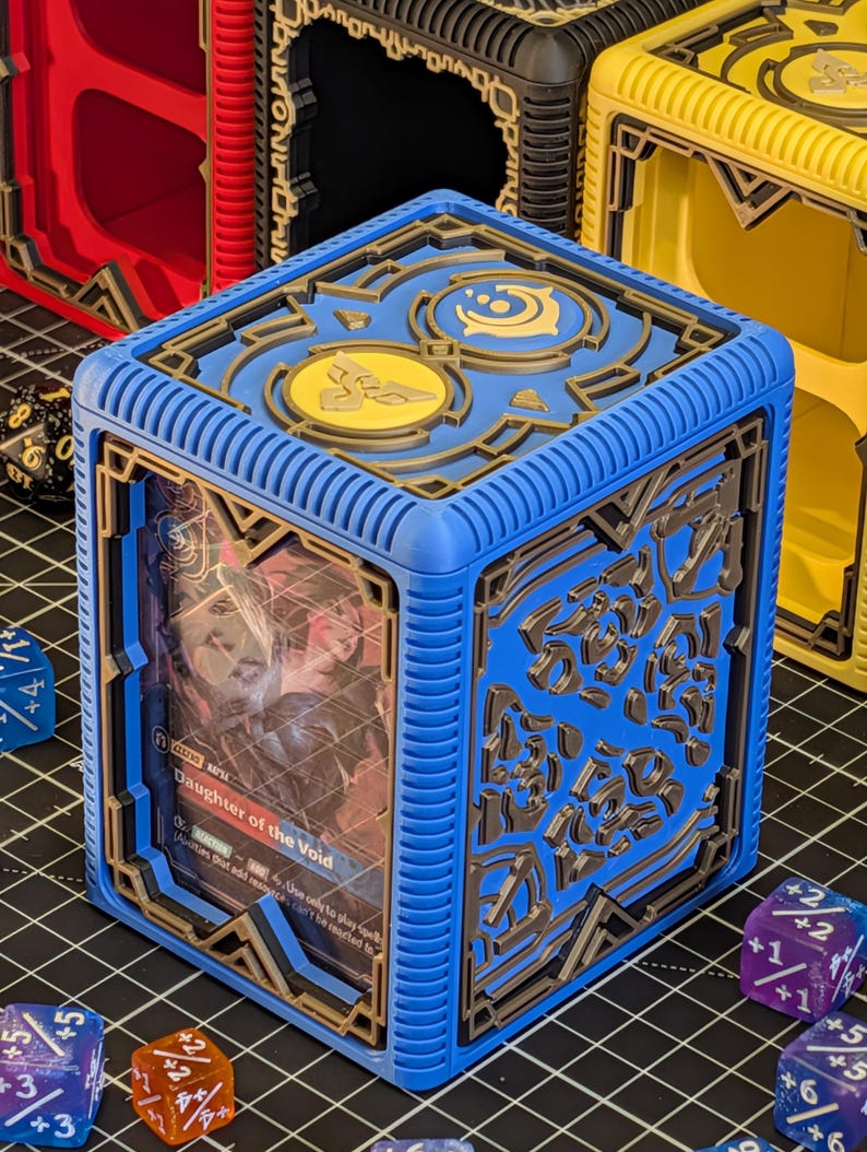 May include: A blue and gold card storage box with intricate geometric designs. The box features a card with the text "Daughter of the Void." Other boxes in red, black, and yellow are visible in the background. Dice are scattered around the box.