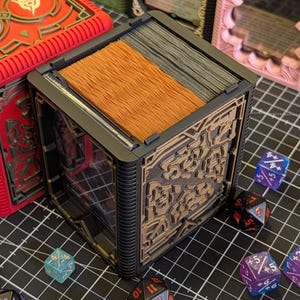 May include: A black and brown card storage box with intricate designs, holding a stack of cards. Several colorful dice are scattered around the box. A red box with a design is visible in the background. The theme is tabletop gaming.