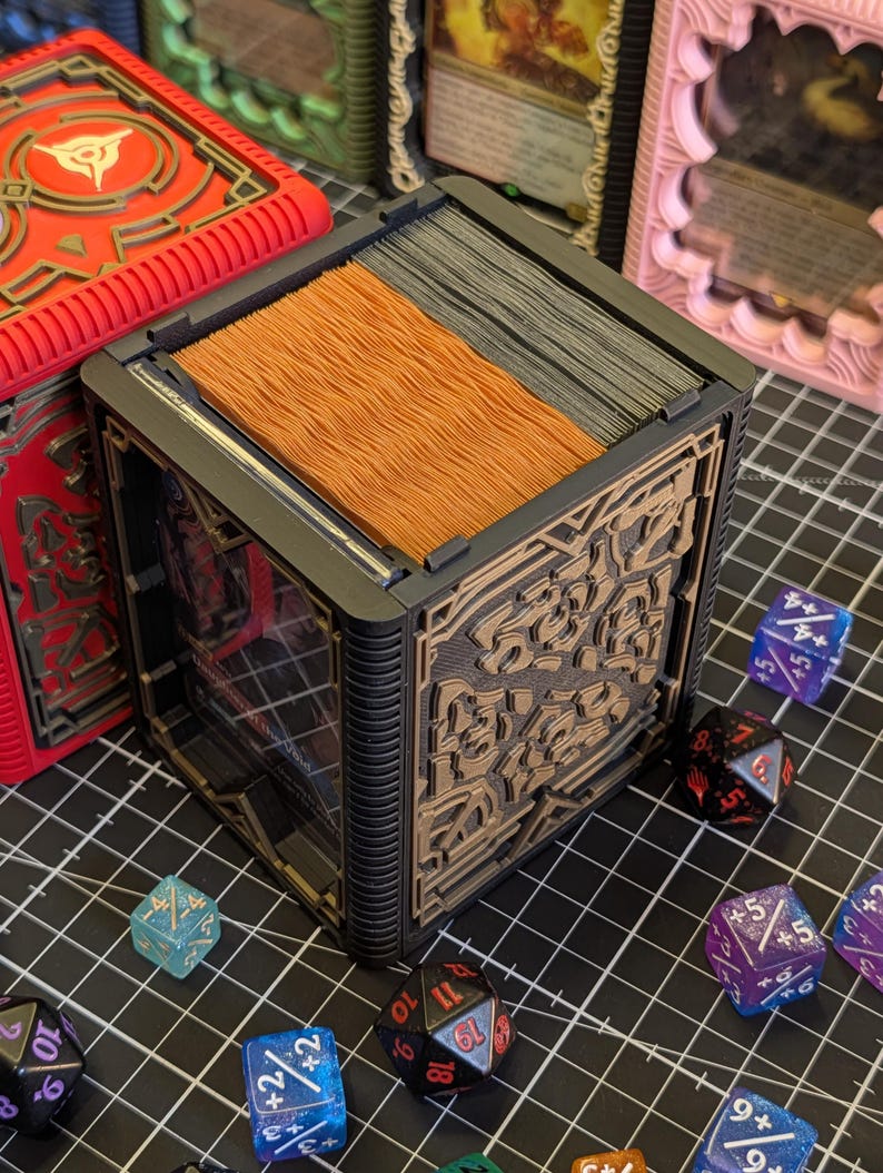 May include: A black and brown card storage box with intricate designs, holding a stack of cards. Several colorful dice are scattered around the box. A red box with a design is visible in the background. The theme is tabletop gaming.