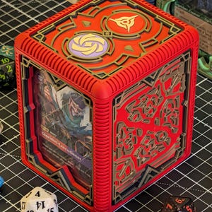 May include: A red, cube-shaped card holder with gold and black accents. The top features two circular designs. A card with the text "Glorious Executioner" is visible inside. Dice are scattered around the holder.