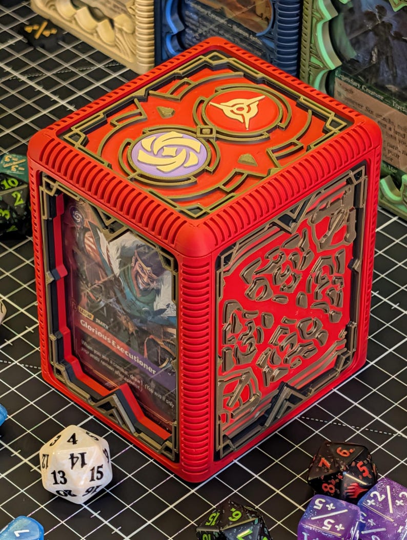 May include: A red, cube-shaped card holder with gold and black accents. The top features two circular designs. A card with the text "Glorious Executioner" is visible inside. Dice are scattered around the holder.