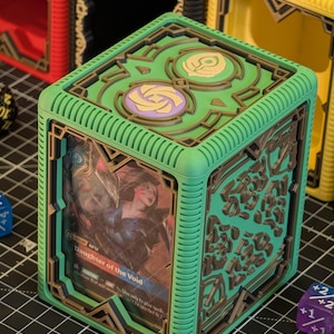 May include: A green card storage box with a brown geometric design. The box features a card with the text "Daughter of the Void". The box is surrounded by dice in various colors.