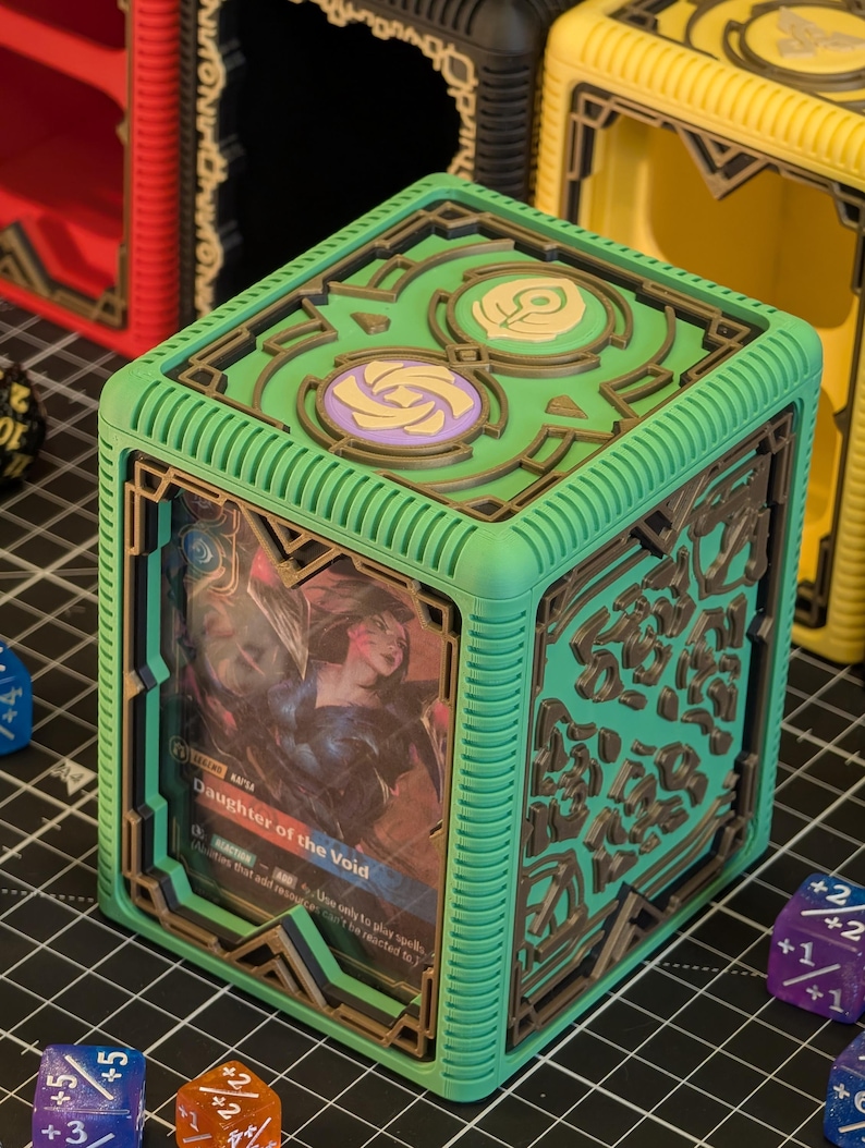 May include: A green card storage box with a brown geometric design. The box features a card with the text "Daughter of the Void". The box is surrounded by dice in various colors.