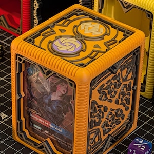May include: Orange and black card storage cube with a card visible that reads "Daughter of the Void". The cube has a decorative design on the sides and top. Other cubes in black and yellow are visible in the background.