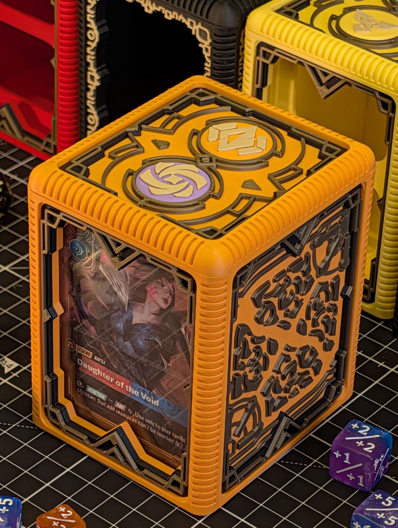 May include: Orange and black card storage cube with a card visible that reads "Daughter of the Void". The cube has a decorative design on the sides and top. Other cubes in black and yellow are visible in the background.