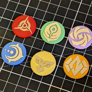 May include: A collection of seven colorful, circular tokens with various beige-colored symbols. The tokens are red, green, purple, blue, yellow, and orange. The tokens are arranged on a black grid.