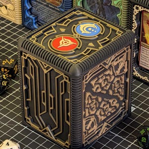 May include: Black and gold cube-shaped card storage box with intricate geometric designs. The top features two circular emblems, one red and one blue. Dice are scattered around the box, suggesting a tabletop gaming theme.