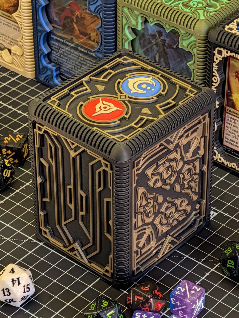 May include: Black and gold cube-shaped card storage box with intricate geometric designs. The top features two circular emblems, one red and one blue. Dice are scattered around the box, suggesting a tabletop gaming theme.