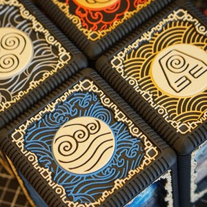 Peut inclure: Gros plan sur quatre boîtes carrées avec des motifs détaillés. Chaque boîte présente un symbole d'élément différent de la série Avatar : feu, eau, terre et air. Les boîtes sont noires avec des accents colorés et des emblèmes circulaires blancs.