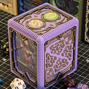 May include: A lilac-colored card storage box with a detailed design, holding a card with the text "Blade Dancer." The box has gold-colored accents and is surrounded by various dice, suggesting a tabletop gaming theme.