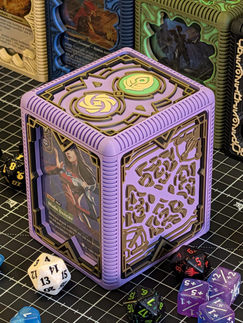May include: A lilac-colored card storage box with a detailed design, holding a card with the text "Blade Dancer." The box has gold-colored accents and is surrounded by various dice, suggesting a tabletop gaming theme.
