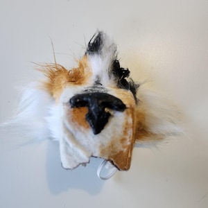 May include: A close-up of a dog's face made of fabric and fur. The face is white, brown, and black with a black nose. The fur is shaggy and the face is slightly tilted to the side.
