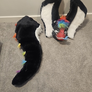 May include: A black and white faux fur creature mask with large ears, a red face, and a rainbow-coloured mohawk. The mask features a silver nose ring and white teeth. A matching black tail with rainbow-coloured spikes runs along the spine.