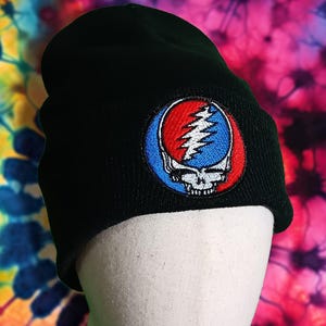 May include: Black beanie hat with a colorful embroidered patch featuring a skull and lightning bolt design in red, blue, and white. The hat is displayed on a mannequin against a tie-dye background.