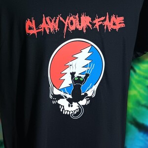 May include: Black t-shirt with the phrase "CLAW YOUR FACE" in red, stylized text. Below the text is a graphic of a skull with a red and blue circle, a lightning bolt, and a black cat with green eyes.