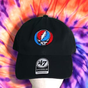 May include: Black baseball cap with a colorful emblem featuring a skull and lightning bolt design. The cap has a curved brim and an adjustable strap. The brand logo "47 CLEAN UP" is visible on the front.