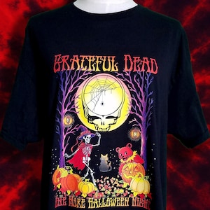 May include: Black t-shirt featuring a Halloween-themed graphic. The design includes a skull, pumpkins, a skeleton, and a bear, with the words "Grateful Dead" and "One More Halloween Night". The artwork is set against a backdrop of a full moon and trees.