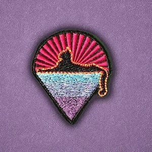 May include: Embroidered patch with a black border, featuring a genie's lamp design. The top section has a pink sunburst pattern, with a black silhouette. The bottom section is a gradient of blue and purple. A gold-colored tassel hangs from the lamp.