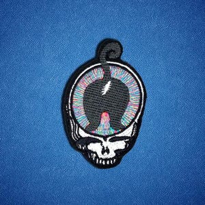 May include: An embroidered patch featuring a black skull silhouette with a white lightning bolt detail at the center. The skull is surrounded by a circular border filled with vibrant, multi-colored radial lines against a dark blue background.