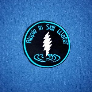 May include: A round, black patch with the words "Ripple in Still Water" in a circle around a white lightning bolt design. The patch has a light blue border and is set against a blue background.