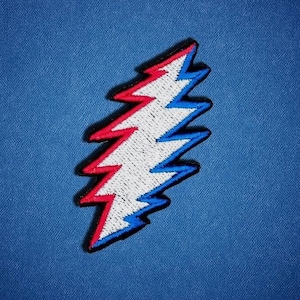 May include: An embroidered patch featuring a jagged lightning bolt design. The bolt is divided into three vertical sections with a red left side, a white center, and a blue right side, all outlined in a thick black border against a blue background.