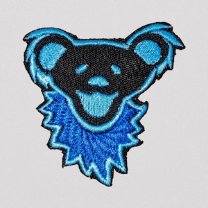 May include: Embroidered patch featuring a stylized bear design. The bear is black with blue accents, including the outline, ears, and a jagged collar. The bear has a smiling face with a tongue sticking out.