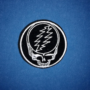 May include: A round, black and white embroidered patch featuring a skull with a lightning bolt design inside a circle. The patch is centered on a blue fabric background. The skull has a detailed, stylized design.