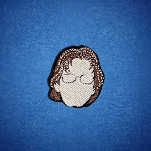 May include: Embroidered patch featuring a stylized face with glasses and brown hair. The face is a light beige color, with brown hair and glasses. The patch is on a blue background.