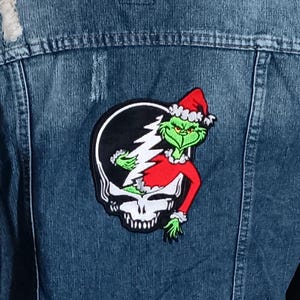 May include: Dark wash denim jacket with distressed details. The back features a patch with a Grinch-inspired design wearing a Santa hat, set against a skull and lightning bolt emblem. The jacket has a classic collar and button closures.