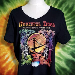 May include: Black V-neck t-shirt featuring a colorful graphic of the Golden Gate Bridge inside a skull, with trees and skeletons. The text "Grateful Dead Celebrating 60 Years 1965-2025" is printed above the graphic in orange and yellow.