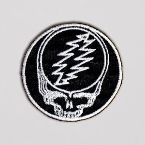 May include: A round, black patch featuring a white skull with a lightning bolt design. The skull has a white outline, and the lightning bolts are in the center. The patch has a white border.