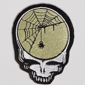 May include: Embroidered patch featuring a skull design with a spider web and spider inside the head. The skull is white with black outlines and a black border. The spider web and spider are black, set against a light green background.