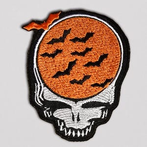May include: Embroidered patch featuring a skull design with an orange circle containing black bat silhouettes. The skull has a white and black color scheme, with a black border. An orange bat is above the circle.