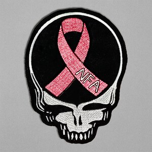 May include: Embroidered patch featuring a white skull with a black background and a pink ribbon. The ribbon has the letters "NFA" embroidered on it. The patch has a white border and is likely for clothing or accessories.