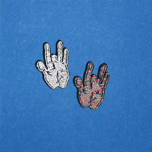 May include: Two embroidered patches in the shape of a hand making the rock on gesture. One patch is white with black stitching, the other is multicolored with a rainbow pattern. The patches are on a blue background.