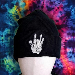 May include: Black knit beanie featuring a white embroidered hand gesture. The beanie is displayed against a colorful tie-dye background with shades of blue, pink, and yellow. The hat is a classic winter accessory.