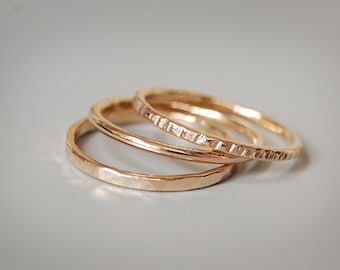 May include: Three gold rings stacked on top of each other. The bottom ring has a hammered texture, the middle ring has a smooth finish, and the top ring has a textured design.
