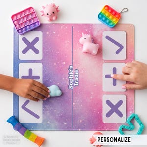 May include: A rectangular game mat with a pink and purple galaxy print featuring the text Sophie's Trades. The mat includes white squares with purple symbols, surrounded by various small silicone fidget toys including a unicorn and axolotl.
