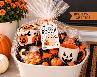 Puede incluir: Una cesta ovalada blanca llena de artículos de Halloween, incluyendo una taza blanca con una cara de calabaza negra, dulces de maíz, una calabaza pequeña y calcetines suaves con patrones de calabaza, todo envuelto en celofán.