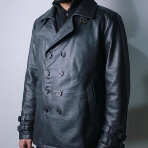 May include: A black leather double-breasted jacket with a notched collar, six buttons, and two side pockets. The jacket has adjustable straps with buckles on the cuffs. The model is wearing a black sweater underneath.