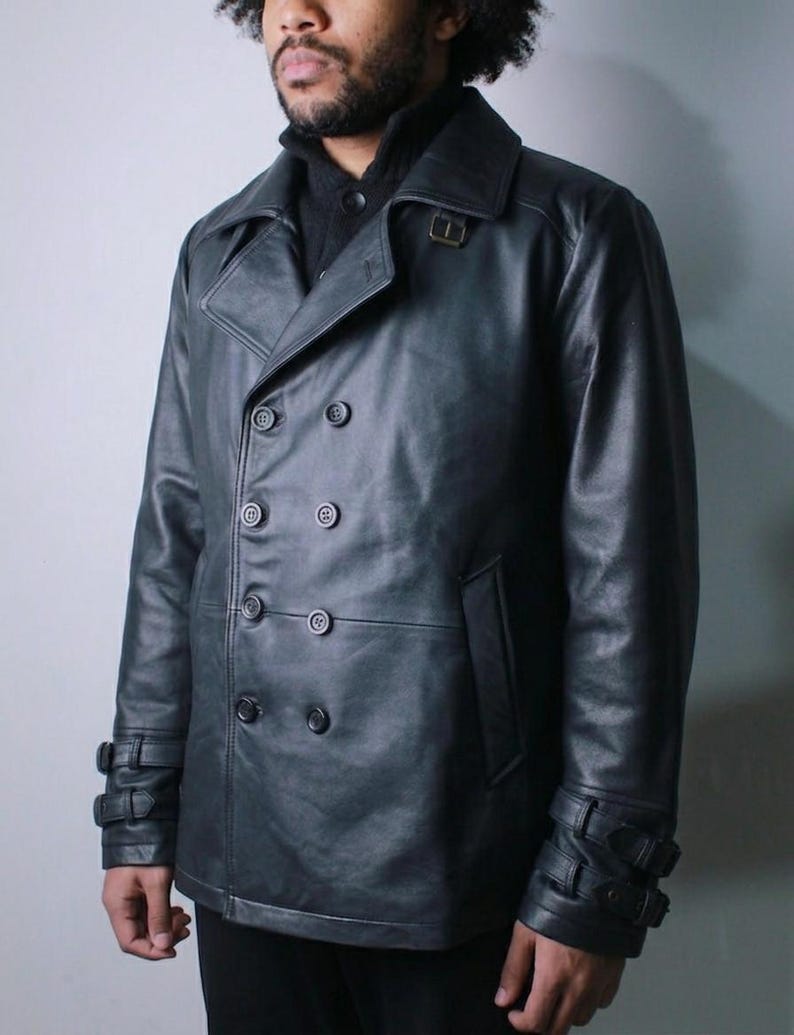 May include: A black leather double-breasted jacket with a notched collar, six buttons, and two side pockets. The jacket has adjustable straps with buckles on the cuffs. The model is wearing a black sweater underneath.